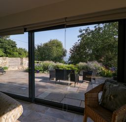 A room with a sofa and wicker chair looking out through large sliding glass doors onto a stone patio with outdoor furniture and garden at Black Cat Bach in Gilwern