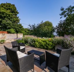 An outdoor patio with woven chairs and table surrounded by stone paving and greenery at Black Cat Bach in Gilwern