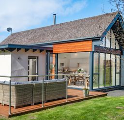 An outdoor patio with wicker seating and cushions next to a house with large glass windows and a wooden deck at Lambs Lodge in Bamburgh