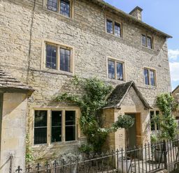 Stone exterior of a multi-story house with windows, a door with a small roof, potted plants, and a metal fence at The Old Post Office in Coln St Aldwyns