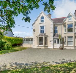 A two story cream-colored house with large windows and a gravel driveway with trees and bushes at Bryn Rhosyn in Nefyn