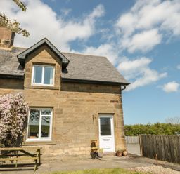 An exterior view of a stone house with two windows a white door a wooden bench and flowering shrub at Appletree in Alnwick