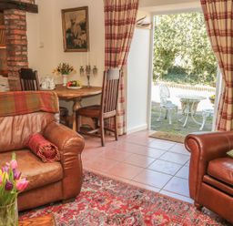 A living room with brown leather sofas a round wooden table with chairs and an open door to a patio at Holly Cottage in Alnwick