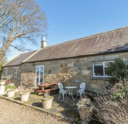 Stone cottage exterior with patio furniture and potted plants under a clear sky at Holly Cottage in Alnwick