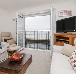 A living room with sofas, armchair, coffee table, television on wooden stand, and open doors with view of the sea at 14 Lyme Mews in Seaton Devon