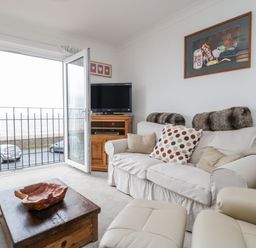 A living room with a sofa, armchair, wooden coffee table, TV on a stand, and an open door to a balcony overlooking the sea at 14 Lyme Mews in Seaton Devon