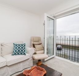 A living room with a white sofa and two blue cushions an armchair and an open door leading to a balcony overlooking the sea at 14 Lyme Mews Seaton Devon