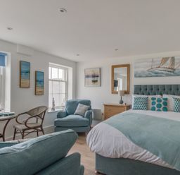 A bedroom with a bed armchairs a small dining table with chairs artworks on walls and windows at 6 The Beaches in Weymouth