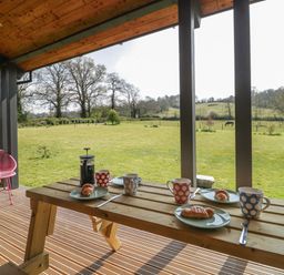 A covered outdoor patio with a wooden picnic table set for breakfast and a grassy field with trees in the background at Bluebell in Sticklinch West Pennard Glastonbury