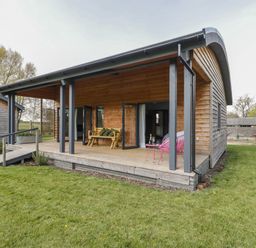 A wooden cabin with a covered porch featuring a bench and pink chairs on a lawn at Bluebell in Sticklinch West Pennard Glastonbury