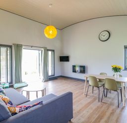 A living room with a grey sofa and colorful cushions a green chair and footstool a dining table with four chairs and yellow flowers at Primrose in Sticklinch West Pennard Glastonbury