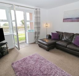 A living room with a leather sofa with purple and white cushions a purple rug a television on a stand a floor lamp and an open door leading outside at Bay View in Benllech