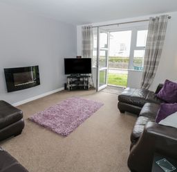 A living room with leather sofas a purple rug a wall-mounted fireplace and a TV near a glass door at Bay View in Benllech