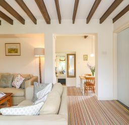 A living room with beige sofas, a wooden coffee table, a floor lamp, and a dining area with a wooden table and chairs at Woodstock Cottage in Birchill near Chardstock