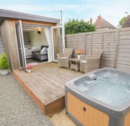A small wooden cabin with an open door leading to a bedroom a wooden deck with two wicker chairs and a table a hot tub next to the deck and a gravel pathway at Alstroemeria Bretforton near Evesham