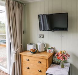 A wooden dresser with a toaster kettle and tea bags a TV on the wall and flowers on a small table near a balcony door at Alstroemeria Bretforton near Evesham