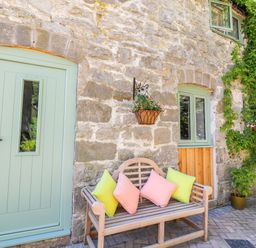 A stone wall with a light green door and windows a wooden bench with colorful cushions and hanging flower baskets at The Old Coach House in Llanarmon Yn Ial