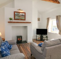 A living room with grey sofas and blue floral cushions a fireplace a TV on a stand and a window with curtains at Macreddin Rock Holiday Cottage Macreddin Aughrim County Wicklow