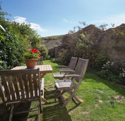 A garden with wooden chairs and a table with a flower pot surrounded by plants and a stone wall at Brock Cottage in West Bay
