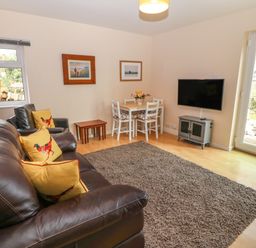 A living room with a brown leather sofa, armchair, small dining table with chairs, wall-mounted TV, and a rug at Plas Marchog Annexe in Rhoscefnhir near Pentraeth