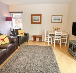 A living room with brown leather sofa and armchair with yellow bird cushions a small dining table with chairs a gray rug and a wall-mounted television at Plas Marchog Annexe in Rhoscefnhir near Pentraeth