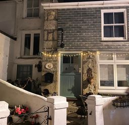 Exterior view of a stone and white house with a green door and windows with string lights above the door at Labour in Vain in St Ives