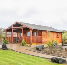 A wooden cabin with a covered porch and a paved path surrounded by grass and rocks at Alken Cabin in Bushmills County Antrim