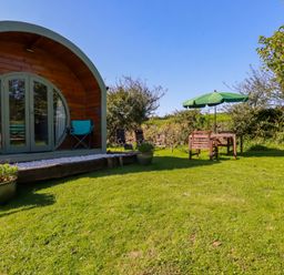 A garden with a wooden pod structure and outdoor seating under a green umbrella at The Lamb Shack in St Teath