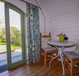 A small dining area with two wooden chairs and a round table next to a glass door with floral curtains at The Lamb Shack in St Teath