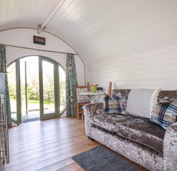 A small living space with a sofa, pillows, a small table with chairs, and a set of large double doors opening to the outside at The Lamb Shack in St Teath