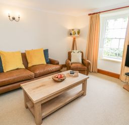 A living room with a brown leather sofa and armchair a wooden coffee table a floor lamp a television and a window with beige curtains at Llety Perygl in Harlech