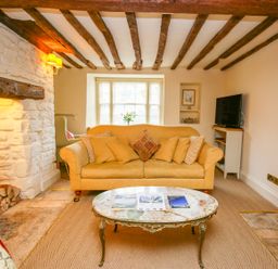 A living room with a yellow sofa a marble coffee table wooden ceiling beams and a stone fireplace at Murton Cottage in Burford