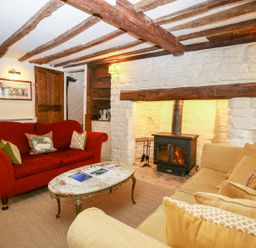 A living room with a red sofa a beige sofa a stone fireplace with a wood stove and wooden ceiling beams at Murton Cottage in Burford