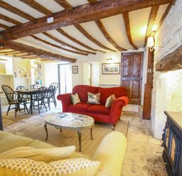 A living room with a red sofa a beige sofa a coffee table a dining table with chairs wooden beams on the ceiling and a stove at Murton Cottage in Burford