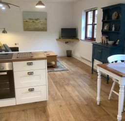 A living area with a kitchen island stove white cabinets wooden floor a blue cabinet a dining table with chairs and a small television on a wall shelf at Mendip Cottage in Shipham