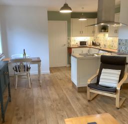 A kitchen and dining area with wooden floor a black armchair and a small dining table with two chairs at Mendip Cottage in Shipham