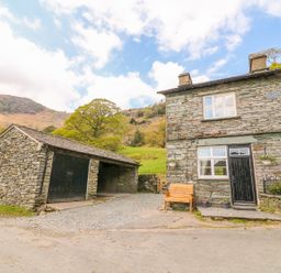 A stone house with a black door and windows next to a stone garage with black doors and a bench outside at Tilberthwaite Farm Cottage in Coniston