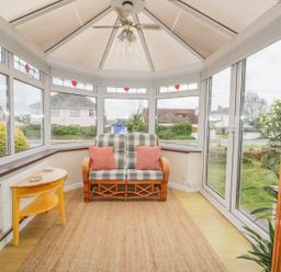 A conservatory with a wicker loveseat with cushions a ceiling fan and a yellow side table at Dune Haven in Goonhavern near Perranporth