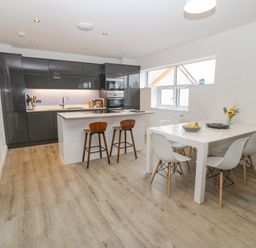 A kitchen with grey cabinets a white island with stools and a white dining table with chairs and flowers at Cysgod Y Capel in Benllech