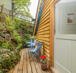 A narrow wooden deck with a half open door beside a small blue table and two blue chairs with cushions and plants nearby at Orchard Loft in Brownston near Modbury