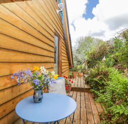 A small wooden deck with a blue round table holding a vase of flowers next to a wooden building and garden at Orchard Loft in Brownston near Modbury