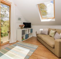 A room with a brown sofa with cushions a white cabinet with a TV a large window and a skylight at Orchard Loft in Brownston near Modbury
