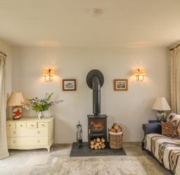 A living room with a wood burning stove flanked by sconces drawers with a lamp and flowers and a couch with cushions and a lamp at The Old Laundry in North Tawton
