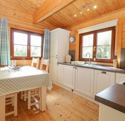 A kitchen with a dining table covered with polka dot tablecloth and white chairs near windows and cabinets at Heron Lodge in Badwell Ash
