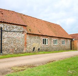 An exterior view of a building with stone walls red tiled roof four white framed windows and a gravel path with grass on the side at Far Barn in Fakenham