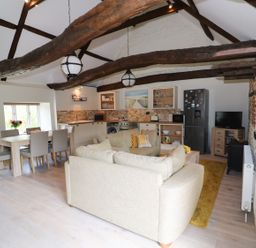 An open-plan living and kitchen area with wooden beams dining table and chairs beige sofas refrigerator and TV at Far Barn in Fakenham