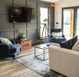 A living room with a television on the wall two blue armchairs a beige sofa a coffee table a wicker basket with books and flowers and sliding glass doors at Meadow View in Meifod Powys