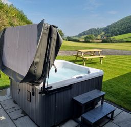 An outdoor hot tub with a partially open cover on a stone patio next to a wooden picnic table on grass at Meadow View in Meifod Powys
