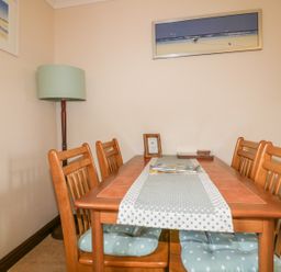 A dining area with a wooden table and six chairs with cushions a table runner a floor lamp and two framed pictures on the walls at Chiffchaff in Padstow