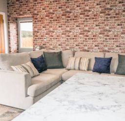 A living room with a beige sectional sofa with cushions a brick wall a wooden door and a marble table at Owl View in Lebberston near Cayton